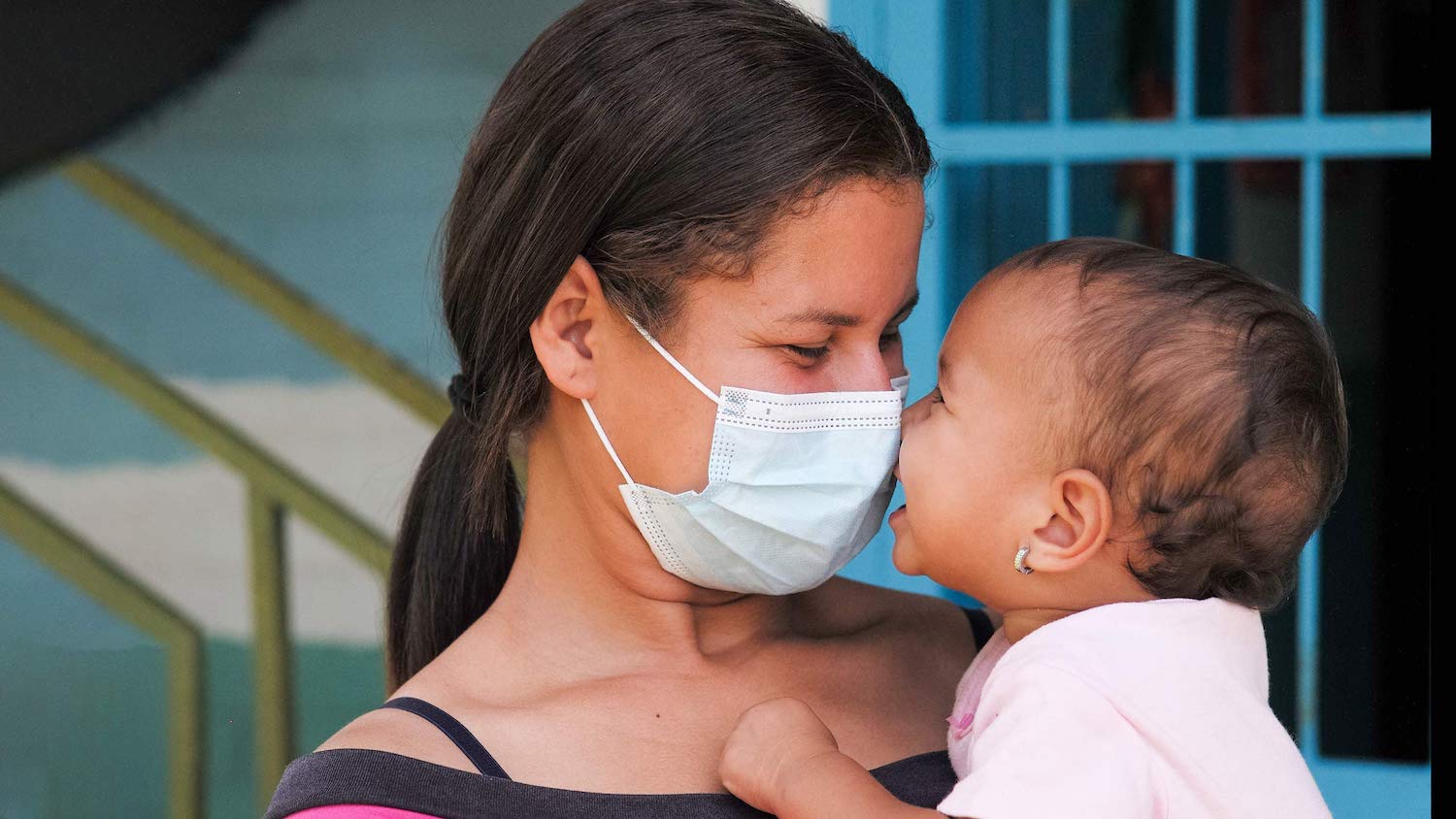 A woman in a mask holds a baby close to her face.