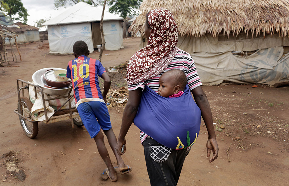 Hawa carries her son as a neighbour pushes a cart with bags of cassava flour, dried fish and nuts for Hawa to sell at the camp’s marketplace. Photo: UN Women/Ryan Brown