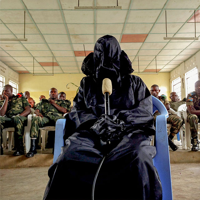 DEMOCRATIC REPUBLIC OF THE CONGO. Minova. 2014. A veiled survivor testifies while 37 Congolese soldiers facing rape charges are seated behind her. Although more than 1,000 sexual assault victims were identified from the 2012 attacks in Minova, only 47 testified. Special care is taken to provide survivors with disguises, curtains, veils, whatever they may need to feel secure when giving their testimony. The women are referred to by numbers instead of by name to maintain their anonymity.  ©Michael Christopher Brown/Magnum Photos