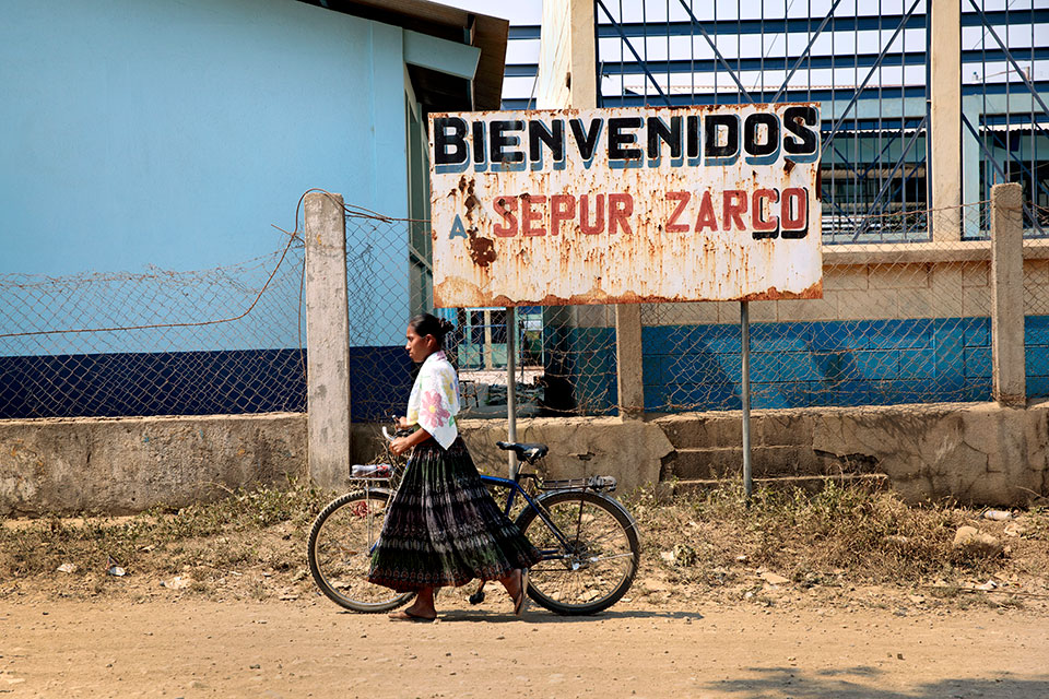 A woman rolls her bike past a sign reading "Bienvenidos Sepur Zarco". Photo: UN Women/Ryan Brown