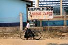 A woman rolls her bike past a sign reading ""Bienvenidos Sepur Zarco"". Photo: UN Women/Ryan Brown
